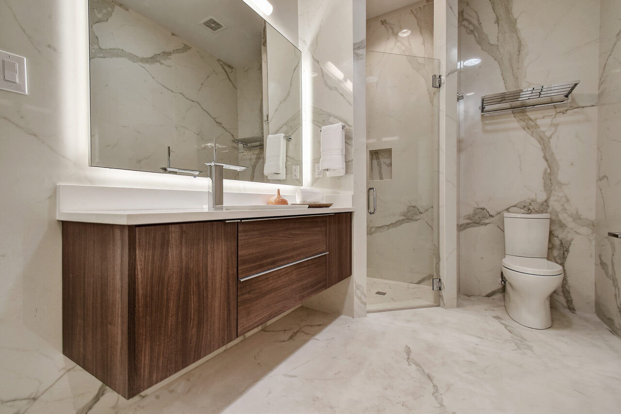 Floating wood bathroom vanity with backlit mirror beside a glass shower